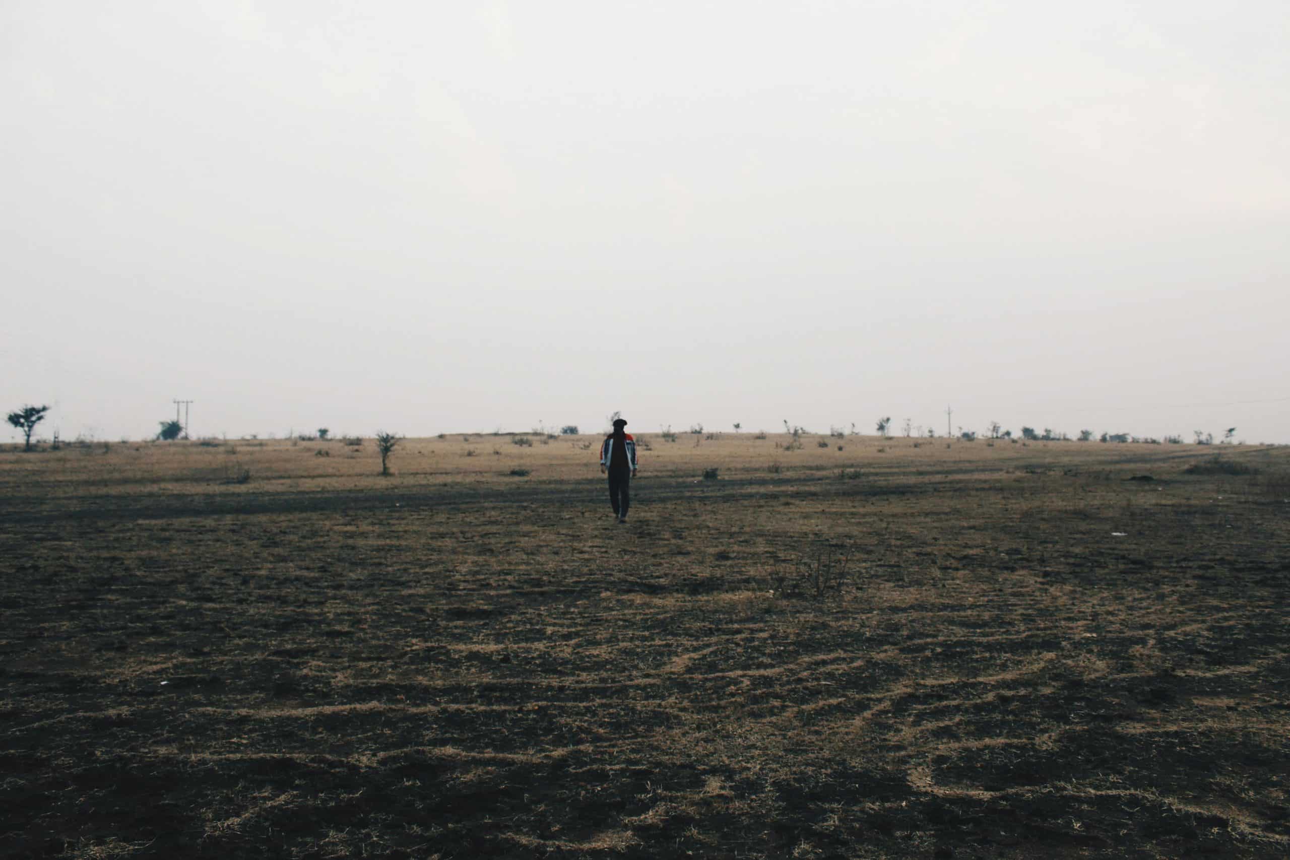 A person walks alone across a vast, barren agricultural field under a cloudy sky.
